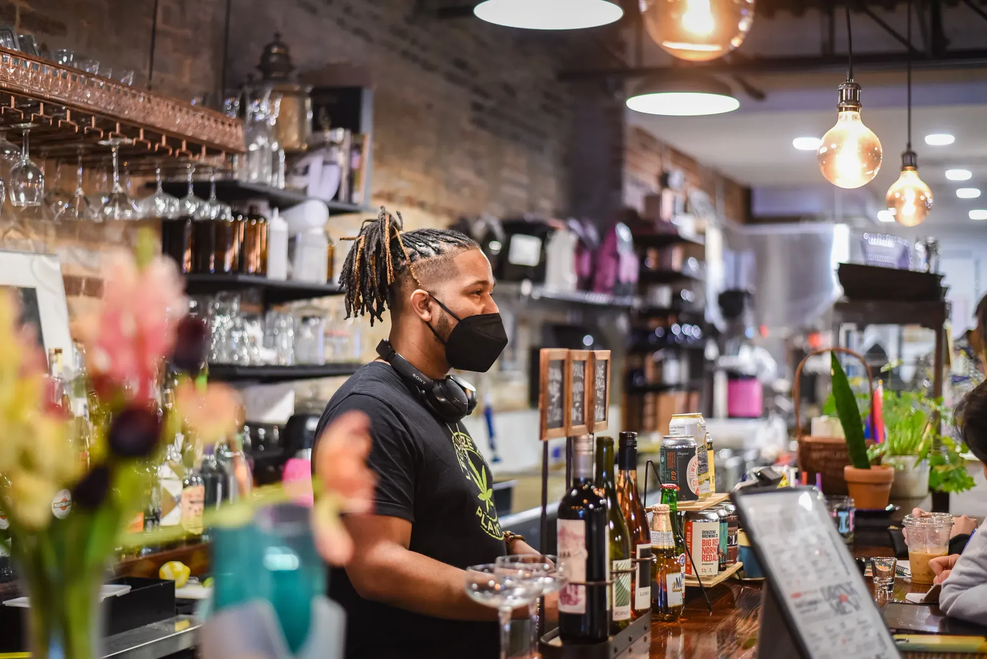 A masked person of colour behind a counter serving customers.