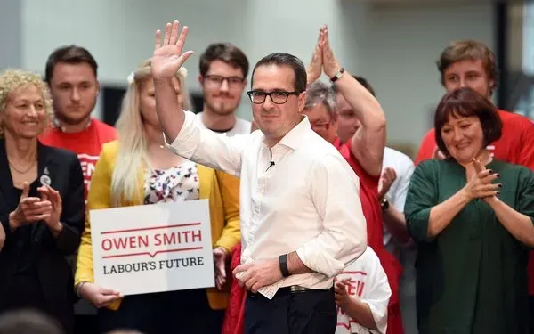 Owen Smith, with close cropped hair, wearing a white shirt, glasses, waving.